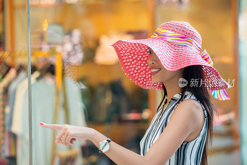 对待自己(购物)。在购物中心的商店橱窗前，一名戴着彩色草帽的亚洲妇女微笑着伸出手指