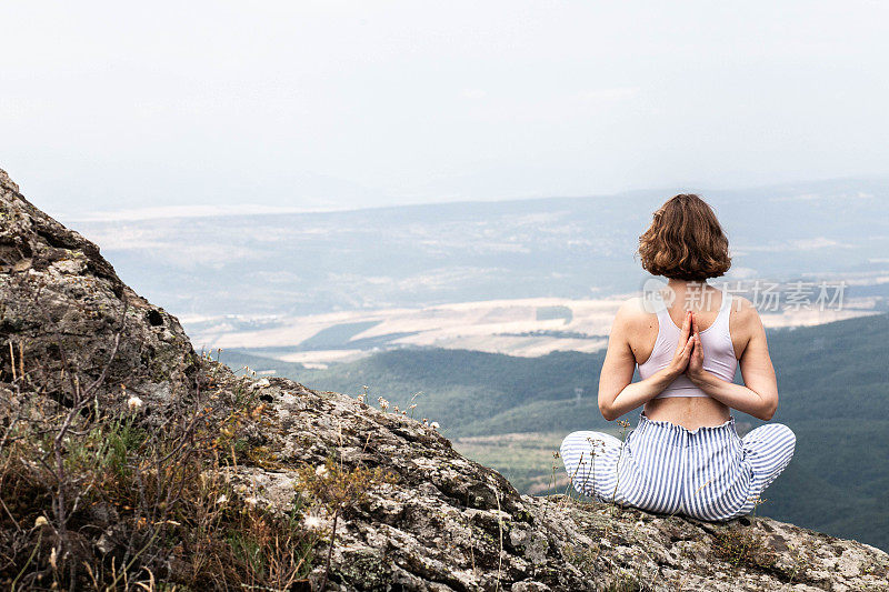 一名身穿白色t恤、留着短发的年轻女子在山外练瑜伽，一名游客背对着镜头坐成莲花状