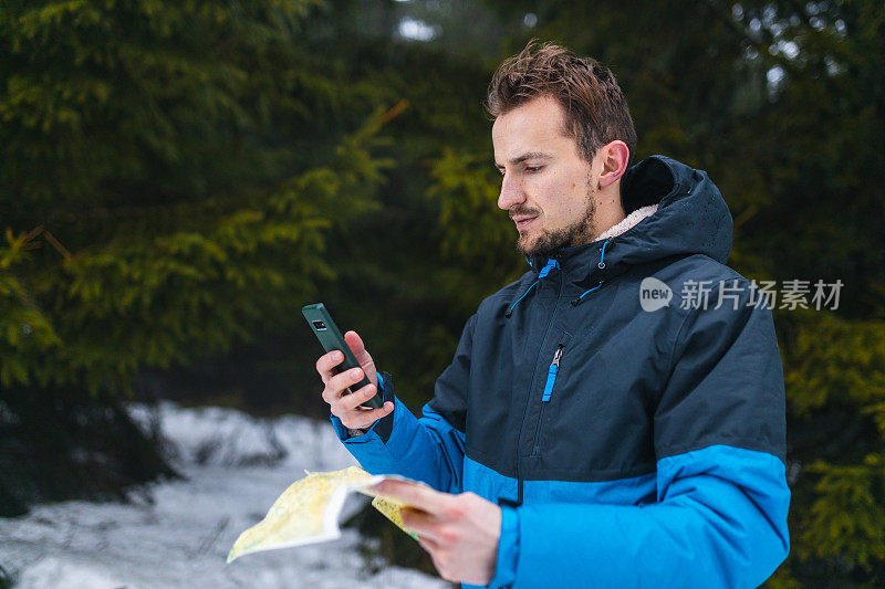 年轻的男性徒步旅行者穿着蓝色夹克，在雾蒙蒙的雪山中迷失了方向，一边看地图，一边看智能手机