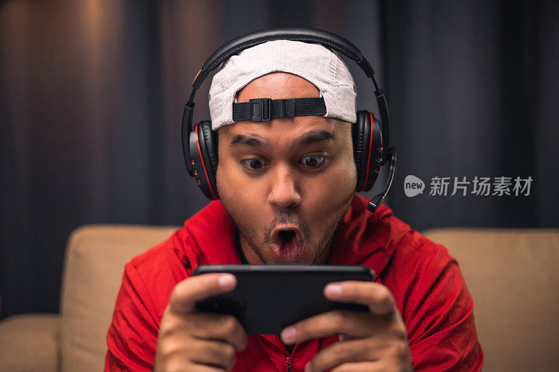 在智能手机上玩游戏。年轻的亚洲帅哥坐在客厅的沙发上拿着手机。印度男子在暗室里戴着耳机在线玩游戏。