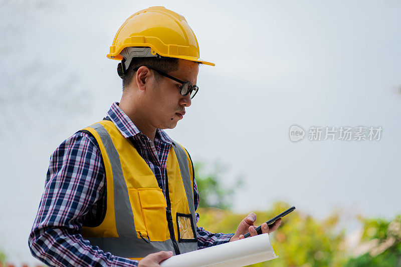 一名年轻的专业工程师戴着防护头盔，拿着智能手机，在一名年轻人的房子建筑工地检查施工文件。