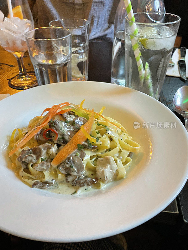 图片的餐厅场景的餐桌设置的奶油鸡肉和蘑菇意大利面条食谱在意大利餐厅，吃出意大利面菜在白色的盘子，喝的杯子，不认识的人在背景