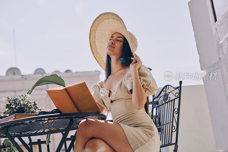 迷人的女人戴着优雅的帽子，拿着书，在阳台上放松