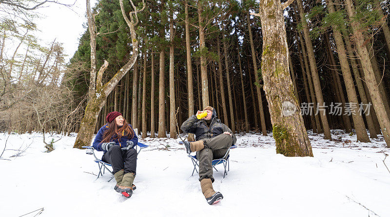 一对情侣在白雪皑皑的森林风景中，在露营椅上放松，分享一瓶热饮