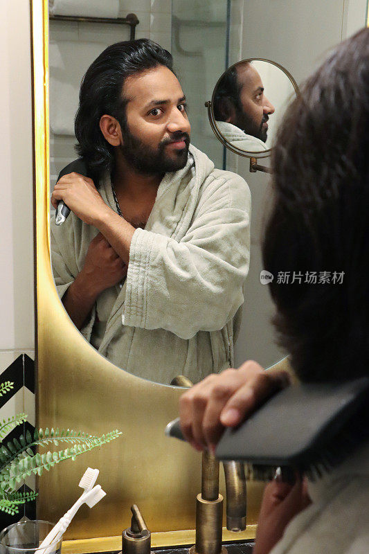 镜子里的特写，印度男人穿着浴袍站在浴室的水池边，用梳子梳理凌乱潮湿的头发，自我护理和修饰的概念