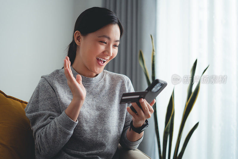 亚洲女性开心兴奋地坐在扶手椅上，用智能手机和信用卡在家网上购物。