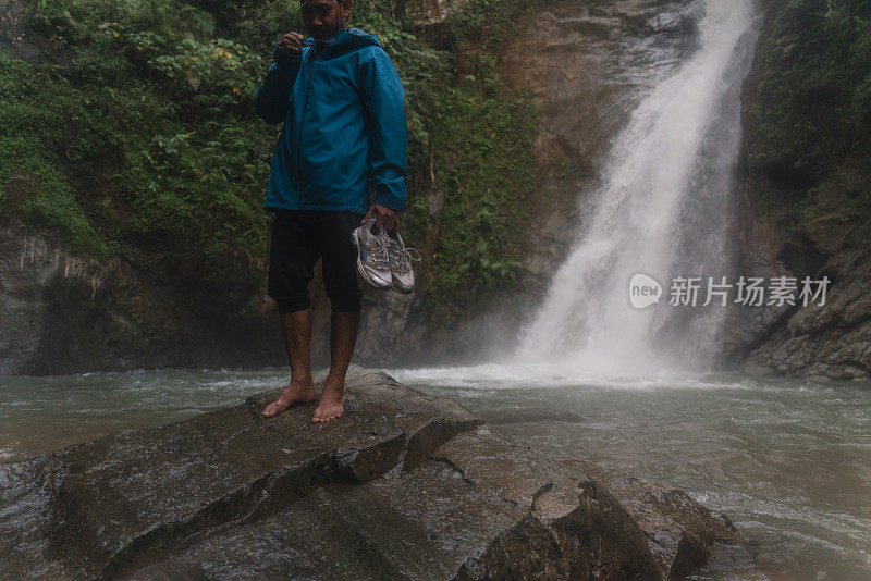 在热带雨林中，一名亚洲男子赤脚走在以瀑布为背景的石头上