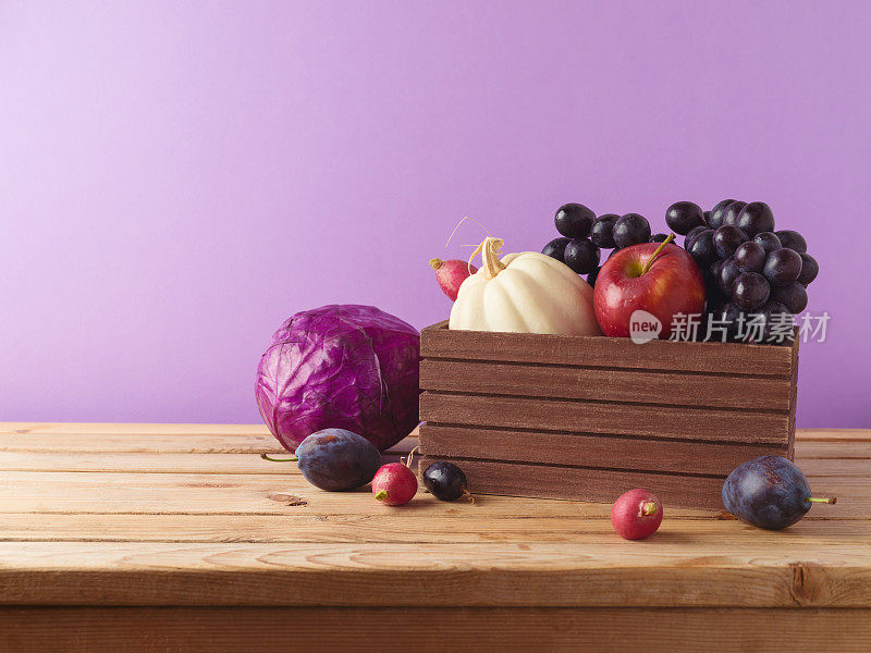 秋收概念。南瓜、苹果、葡萄和蔬菜放在紫色背景的桌子上的木箱里