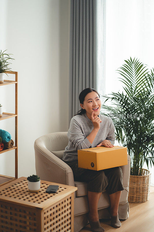 亚洲女人拿着纸盒，开心兴奋地坐在家里客厅的扶手椅上。