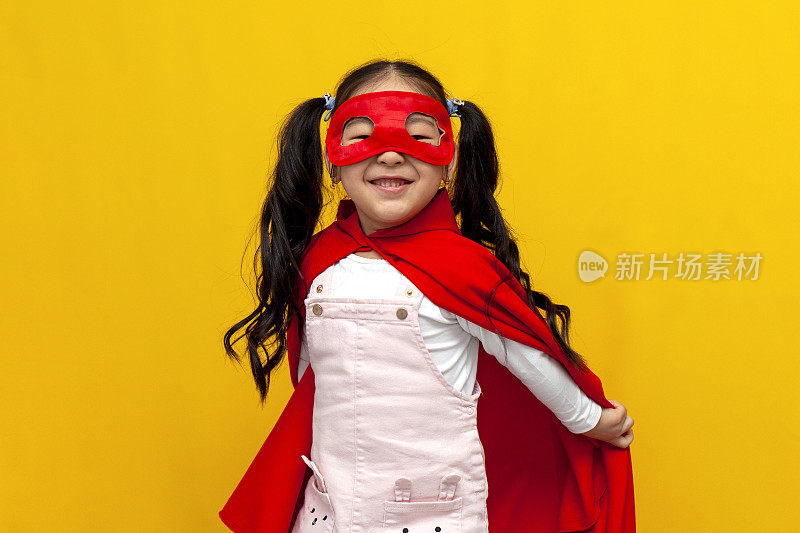 穿着超人服装和面具的亚洲小女孩微笑着，黄色背景上穿着超级英雄斗篷的韩国孩子