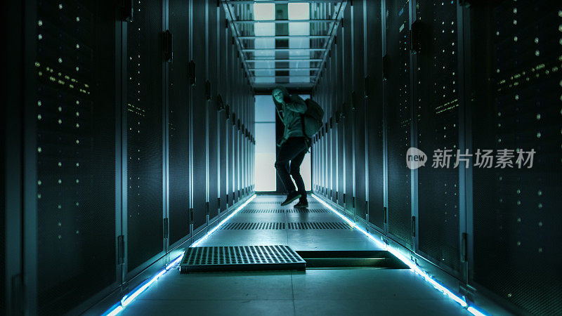 一个穿着连帽衫的蒙面黑客在公司数据中心跑开，拿着笔记本电脑向后看。