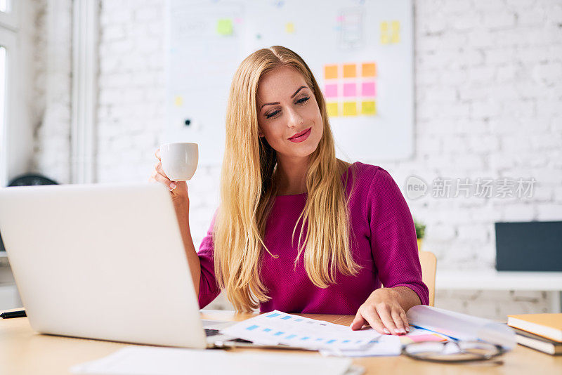 迷人的女商人一边喝咖啡一边在她现代的办公室里做文书工作