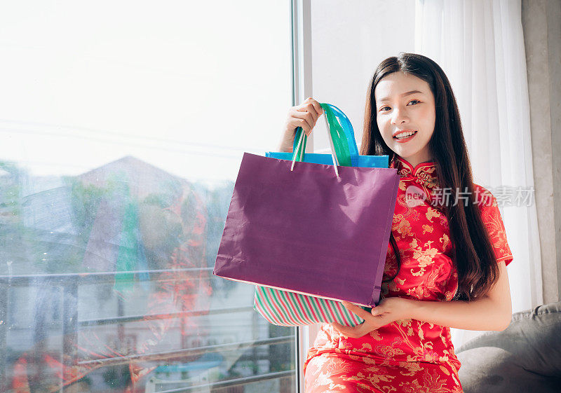购物亚洲女人中国服装，旗袍，旗袍春节和手提袋坐在靠近窗户的清洁白色房间，复印空间。
