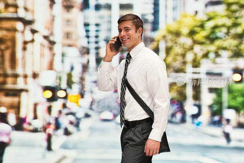 背景模糊的年轻男性商人，在户外背着肩包，拿着包，使用手机