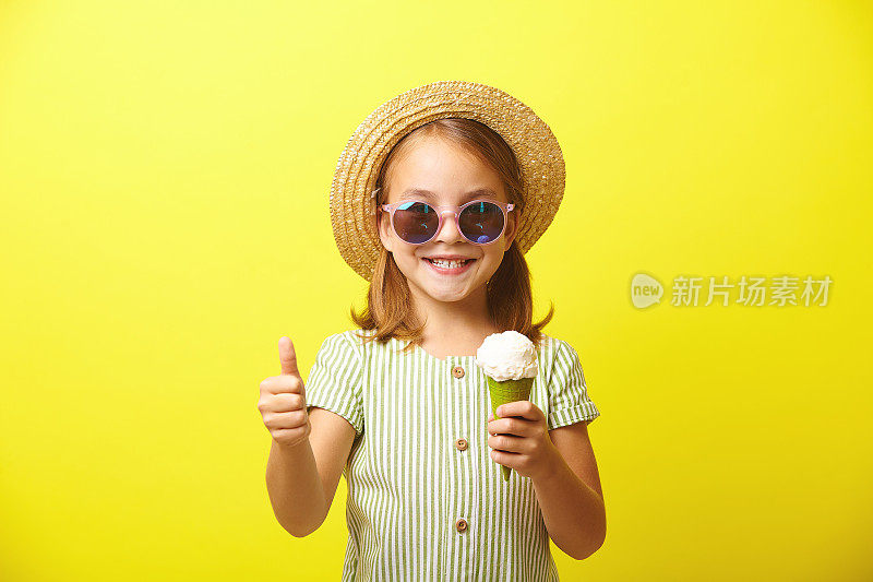 一个快乐的小女孩手里拿着一个漂亮的冰淇淋，翘着大拇指，露出快乐的表情，穿着一件夏装，戴着一顶草帽和太阳镜，站在黄色的孤岛上。