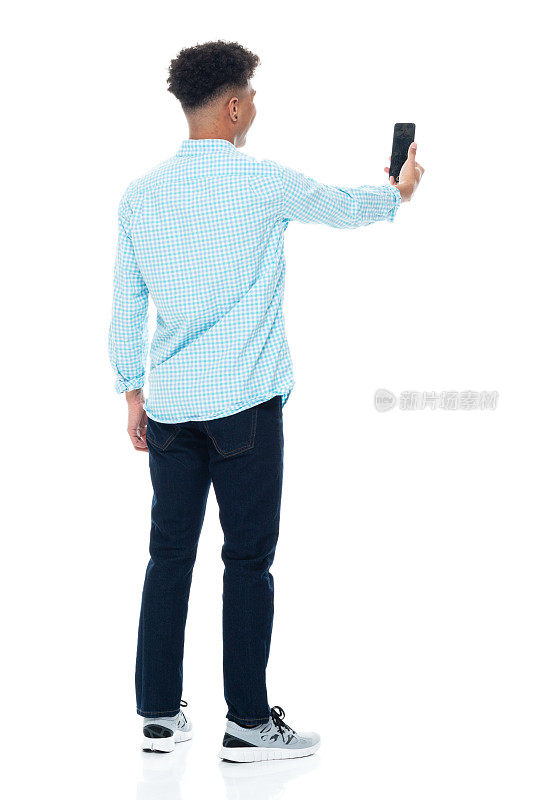非裔美国男孩穿着衬衫，用智能手机站在白色背景前拍照