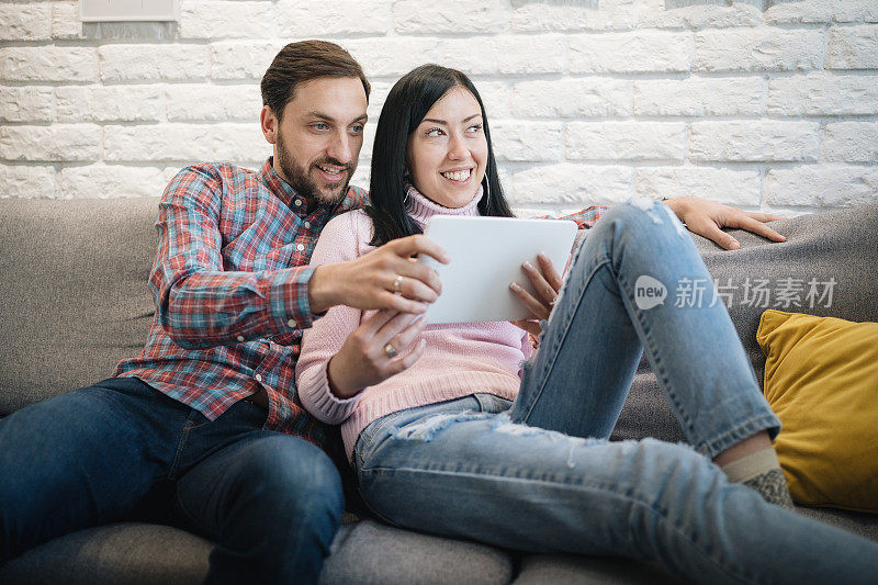 这是一对愉快的年轻夫妇在家里的沙发上休息时一起浏览电子平板电脑的照片