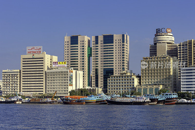 阿拉伯联合酋长国迪拜——清晨阳光下，几艘传统的阿拉伯单桅帆船并排停泊在迪拜河德拉一侧的码头上;背景中，现代办公和住宅建筑。