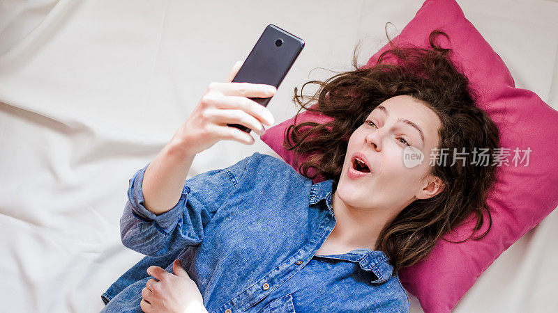 开心惊喜的女人躺在床上用手机和朋友在社交网络上聊天。室内肖像的惊奇时尚女孩在休闲服装拘留牢房，看视频