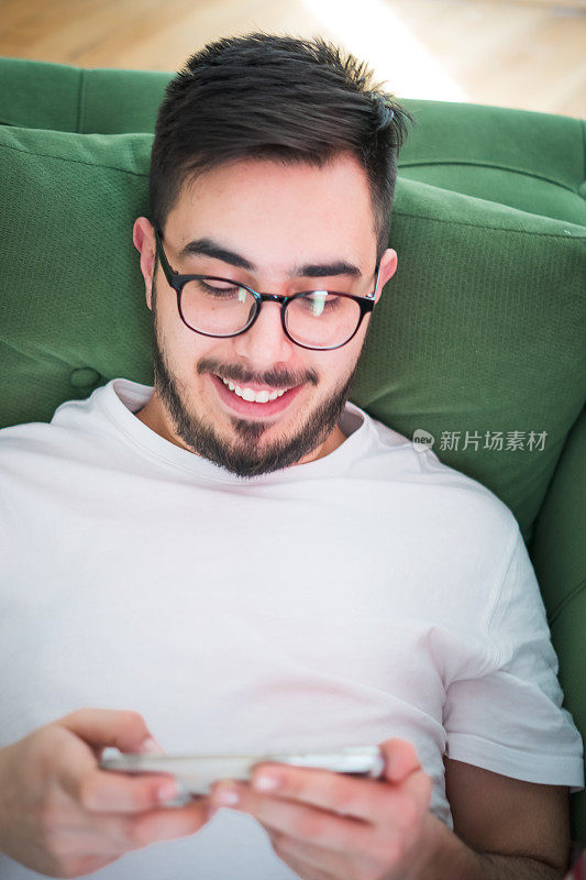 一个年轻人躺在家里的沙发上，用着他的智能手机。