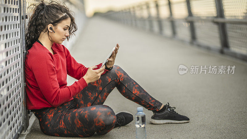 年轻的运动女性在户外艰苦训练后休息，听音乐，使用数码平板电脑和喝水