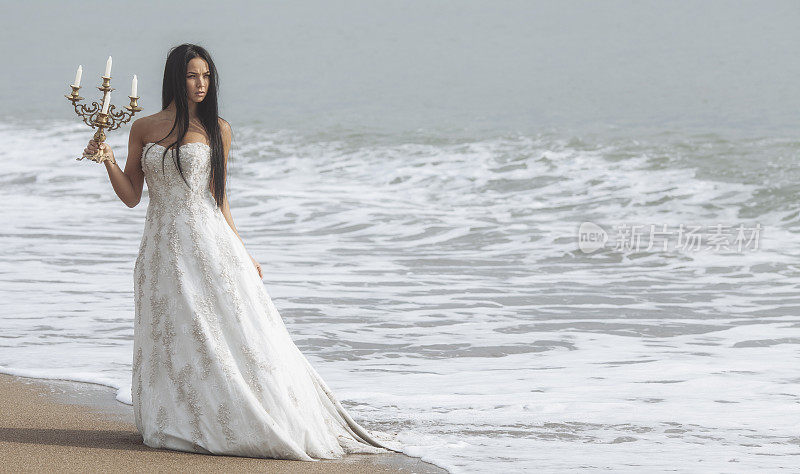 愤怒的年轻女子穿着婚纱，手拿烛台站在海边。