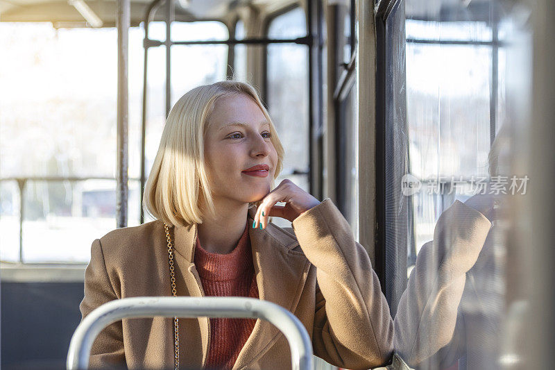 一个年轻的女人在公共汽车上微笑，看着窗外美丽的年轻女人乘公共汽车上班。公共汽车或火车上的乘客，技术生活方式，交通和旅行概念。