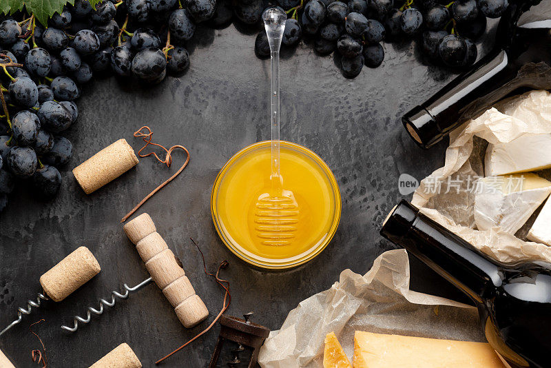 蜂蜜装在玻璃罐里，蜂蜜勺放在深色石板上，葡萄酒奶酪开塞钻装在黑色多汁葡萄框架内。在深色混凝土背景下品尝红酒芝士小吃