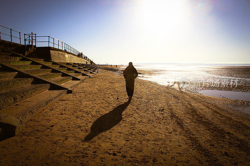 这是一个令人惊叹的照片，一位男性在一个寒冷的冬天早晨穿着冬装走在海滩上。当雄性走向明亮的太阳时，它的影子在沙滩上清晰可见。
