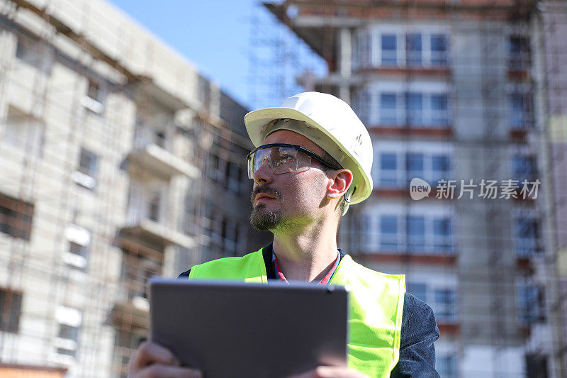 男建筑师在工地使用数码平板电脑，他穿着防护背心和头盔