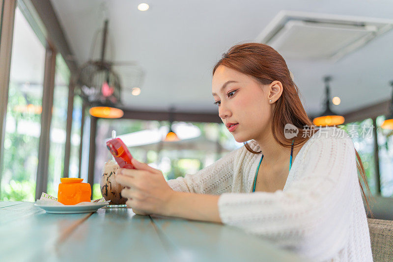 年轻的亚洲女子在海滩咖啡馆一边喝着冰拿铁和橘子蛋糕一边玩智能手机。穿着沙滩装的女性数字游民在度假氛围中使用手机。穿着沙滩装的亚洲女人在咖啡馆里放松。