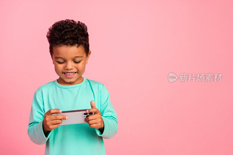 照片肖像迷人的小男孩玩手机游戏有乐趣穿时髦的海蓝宝石服装孤立在粉红色的背景