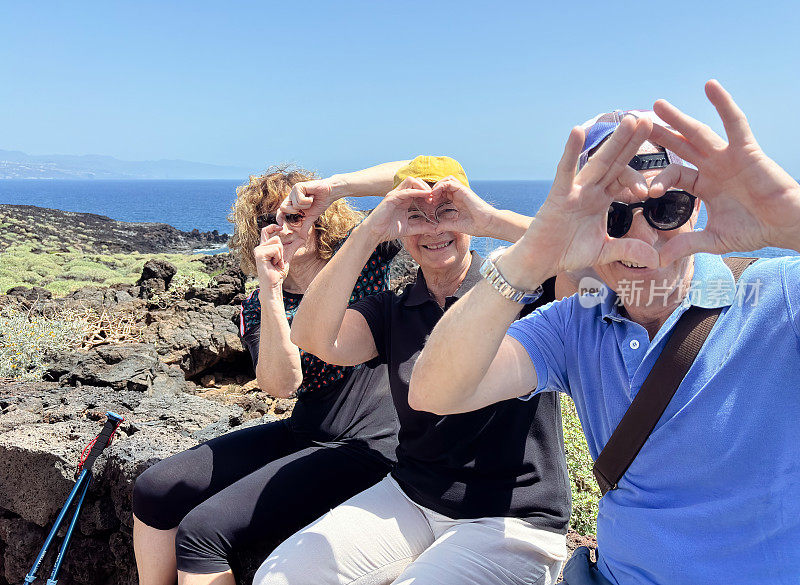 一群英俊的人在海上户外远足，三个白种人朋友坐在悬崖上用手做成心形，微笑着享受自由和假期，退休后的健康活动