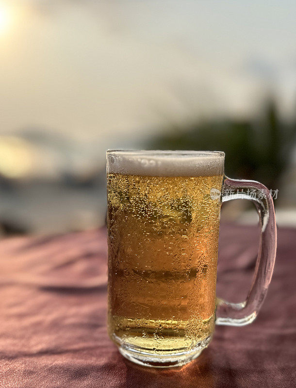 啤酒在玻璃大酒杯的形象覆盖在凝结与起泡的气泡形成泡沫的啤酒顶部的泡沫，马克杯品脱玻璃杯在沙滩酒吧户外桌子与桌布，沙滩在日落，阳光明媚的天空，重点在前景