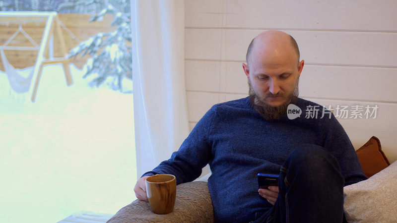 一个留着胡子、秃头的野蛮男人坐在家里的沙发上，喝着早上的咖啡，用智能手机看新闻，他旁边有一台留声机，里面放着黑胶唱片播放音乐。