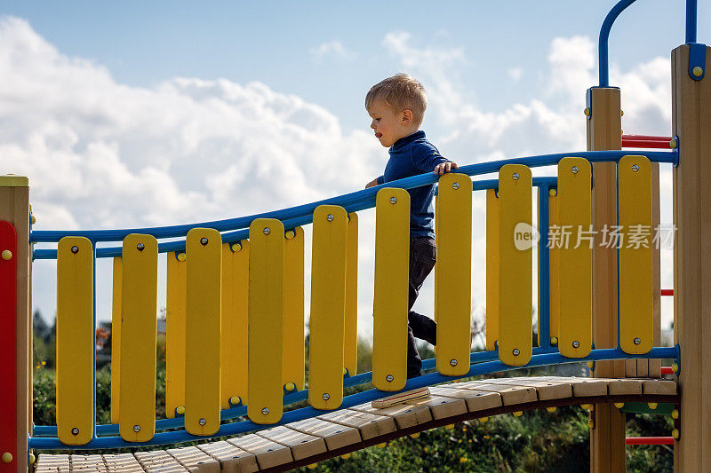 小男孩聚精会神地沿着操场上的弧形桥走着。弯弯的黄色栅栏，以白云为背景的浅蓝色天空
