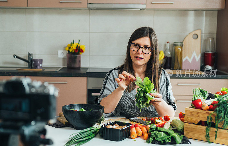 一名妇女坐在厨房的桌子上，拿着生菜，为她的食物频道拍摄视频