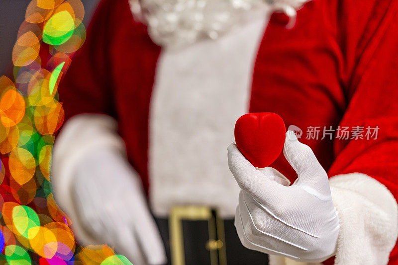 圣诞老人拿着一个心形首饰盒，背景是来自圣诞树的散焦