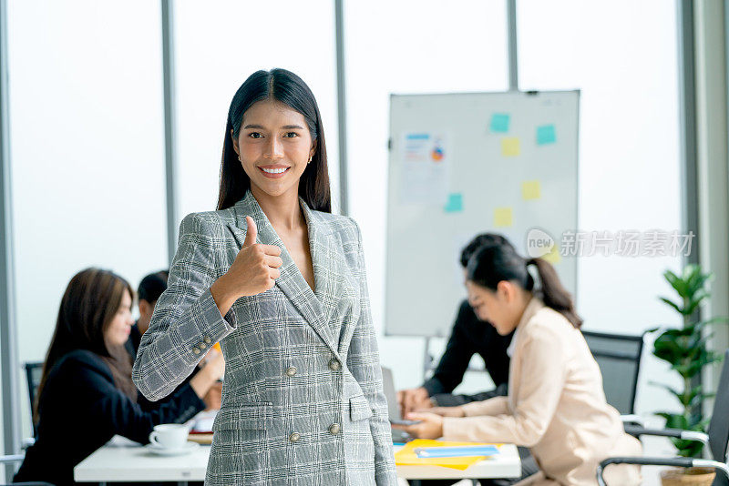 亚洲商业女性的肖像站在镜头前，微笑着向其他同事竖起大拇指，在办公室后面讨论。