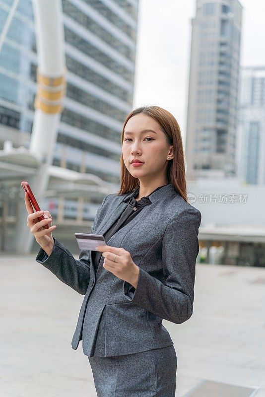 一名年轻的亚洲女商人穿着灰色的办公套装，在商业区用她的智能手机使用信用卡进行在线交易，她微笑着自信