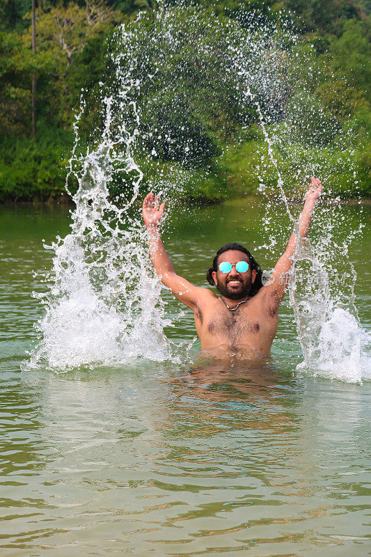 一名印度男子戴着有色的镜面太阳镜，站在齐腰深的泻湖平静的水中，笑着举起手臂，溅起水花，热带岛屿天堂，林地和棕榈树的景色，重点在前景