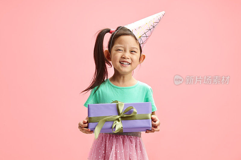 亚裔小女孩戴着派对帽，穿着节日服装，抱着她的生日礼物，她的梦想终于实现了，她快乐地站在粉红色的孤立中。