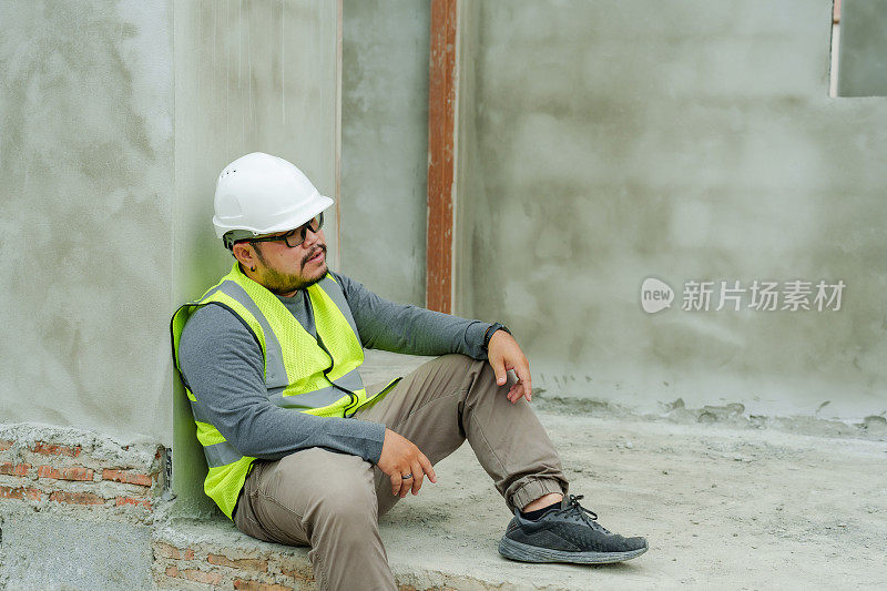 一名穿着高能见度背心的男子坐在一个建筑平台上休息，戴着安全帽，看上去若有所思。建筑师和工程师监督房屋建设。