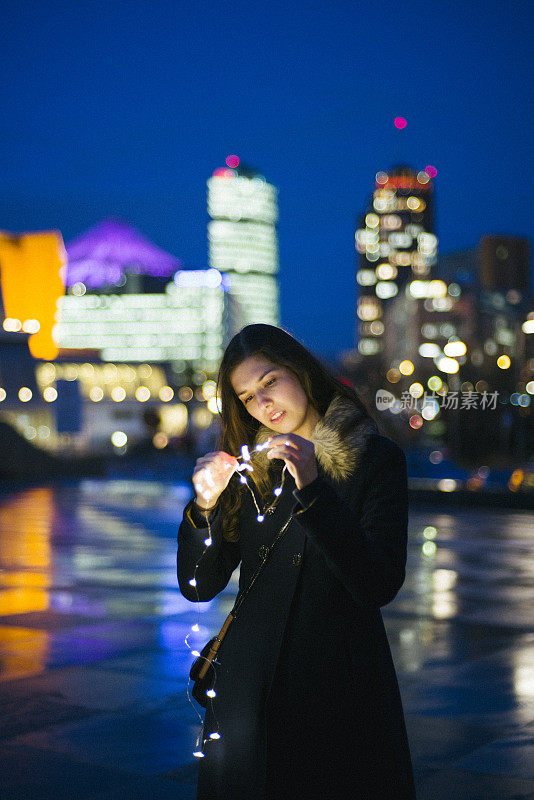 年轻女子站在灯火通明的城市前，手持串灯