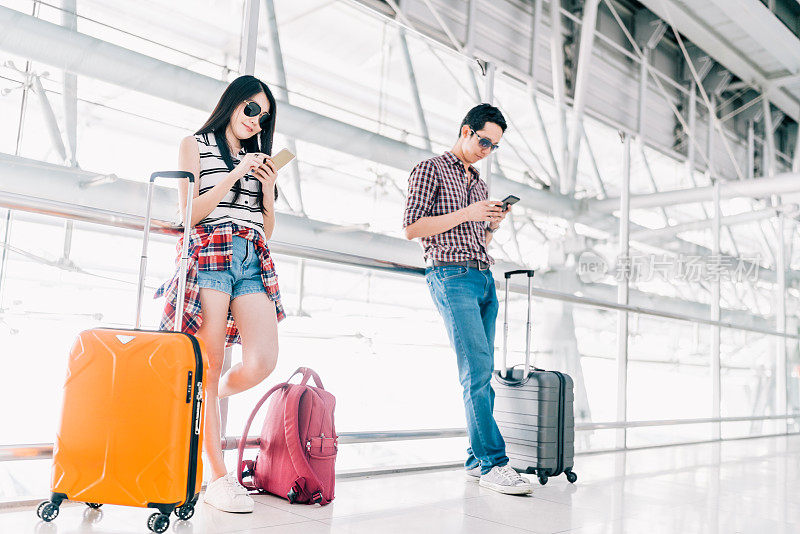年轻的亚洲男性和女性一起在机场用智能手机检查航班或在线值机，并带着行李。航空旅行，暑假，或手机应用技术概念