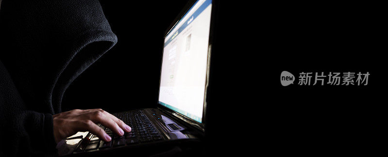 身份不明的黑兜帽黑客使用笔记本电脑。选择性聚焦，在全景黑色背景与复制空间