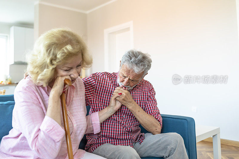 悲伤的退休妻子拄着拐杖坐在客厅里，旁边是老丈夫。老夫妇在家里手拉手看着对方