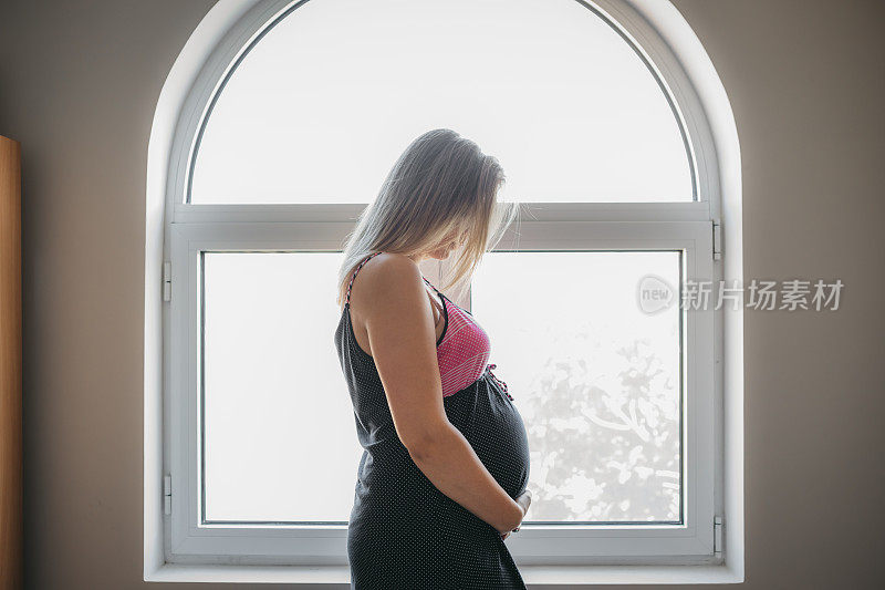 年轻快乐的孕妇在家里的窗户边摆姿势，摸着自己的肚子