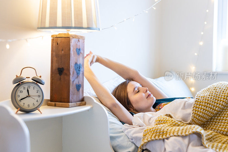 疲惫的金发年轻女子在床上晚了早上，伸展手臂与闹钟在前景时睡过头