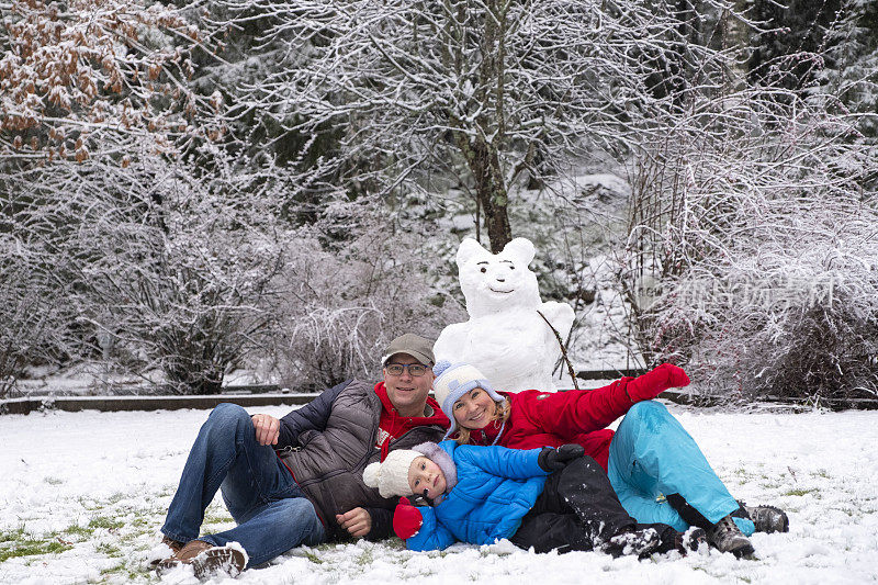 一个白人家庭带着一个5岁的女孩，在雪地里堆了一个雪人，所有人都躺在一起，看着摄像机。